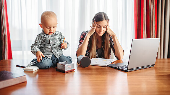 Erwerbsarbeit und private Care-Arbeit müssen zusammen gedacht werden Eine sichtlich gestresste Mutter versucht zu Hause am Laptop zu arbeiten, während neben ihr auf dem Schreibtisch ein Kleinkind sitzt und mit Büroutensilien spielt.