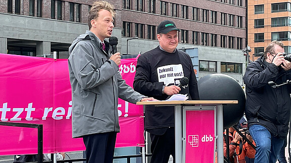 210817 dbb jugend GDL Jugend Zwei junge Männer stehen auf einem Podest auf einer Demonstration, der linke der beiden, Florian Schütz, spricht in ein Mikrofon.