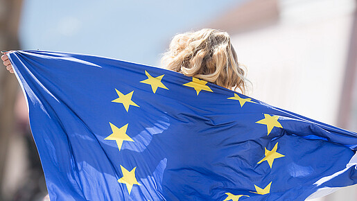 Das Foto zeigt ein Mädchen, das eine EU-Fahne als Umgang nutzt. Die Sterne sind zu erkennen, über dem Rand der Fahne ihre Haare.