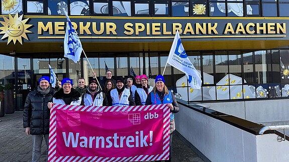 Eine Gruppe Menschen demonstriert vor einem großen Gebäude mit der Aufschrift "Merkur Spielbank Aachen"