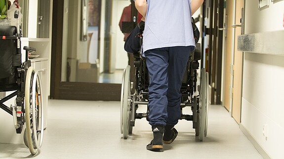 Pflege Eine Pflegekraft, von hinten zu sehen, schiebt einen älterne Menschen im Rollstuhl über einen Flur .