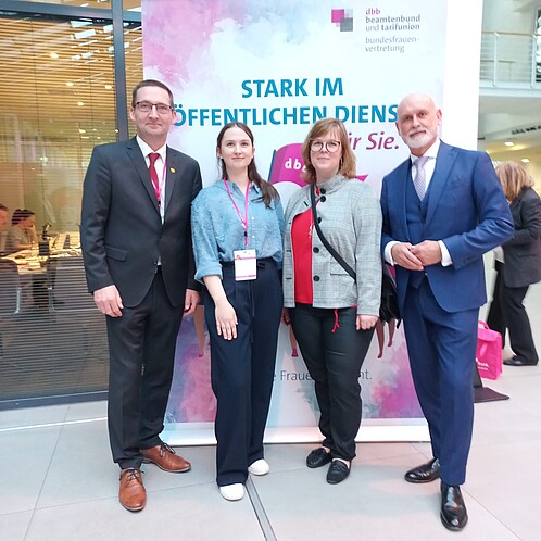 Zwei Frauen und Zwei Männer in formaler Kleidung stehen vor einem Aufsteller. 