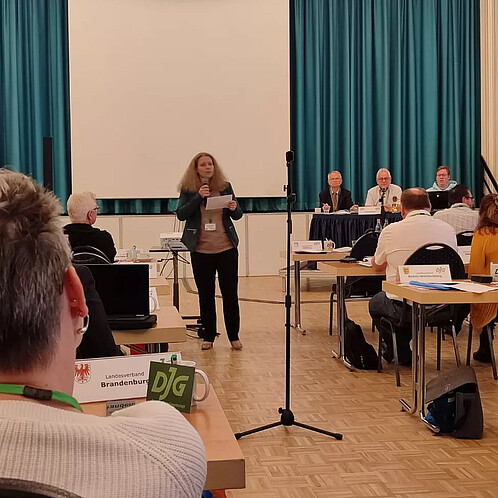 Bianca Korbanek auf dem DJG Bundesgewerkschaftstag 2023 Eine Frau steht in der Mitte eines Seminarraums und spricht vor den anderen Anwesenden in ein Mikrofon