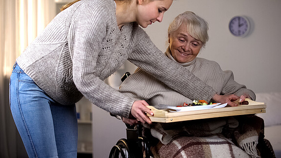 Das Foto zeigt eine Frau, die sich um eine Angehörige kümmert. Sie serviert Essen auf ein Tablett am Rollstuhl.