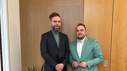 Zwei Männer in Anzügen stehen vor einer Holzwand und blicken in die Kamera