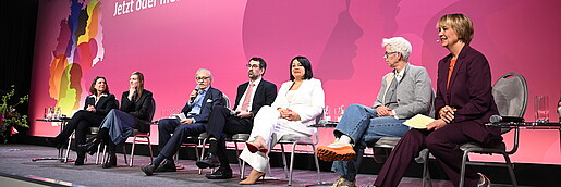 Menschen sitzen auf Stühlen auf einer Bühne und diskutieren miteinander. 
