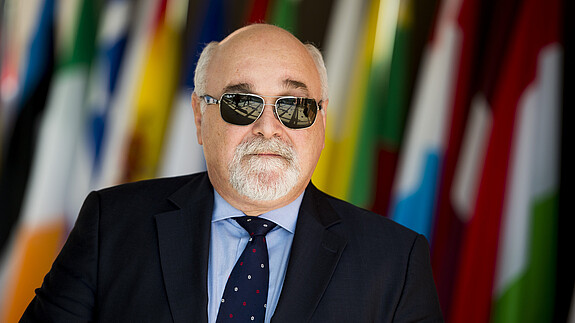 Ioannis Vardakastanis im Portrait, im Hintergrund Fahnen der EU-Mitgliedstaaten