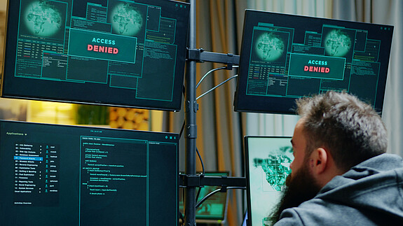 Ein Mann sitzt vor mehreren Computer-Monitoren, auf denen "Zugriff verweigert" zu lesen ist.